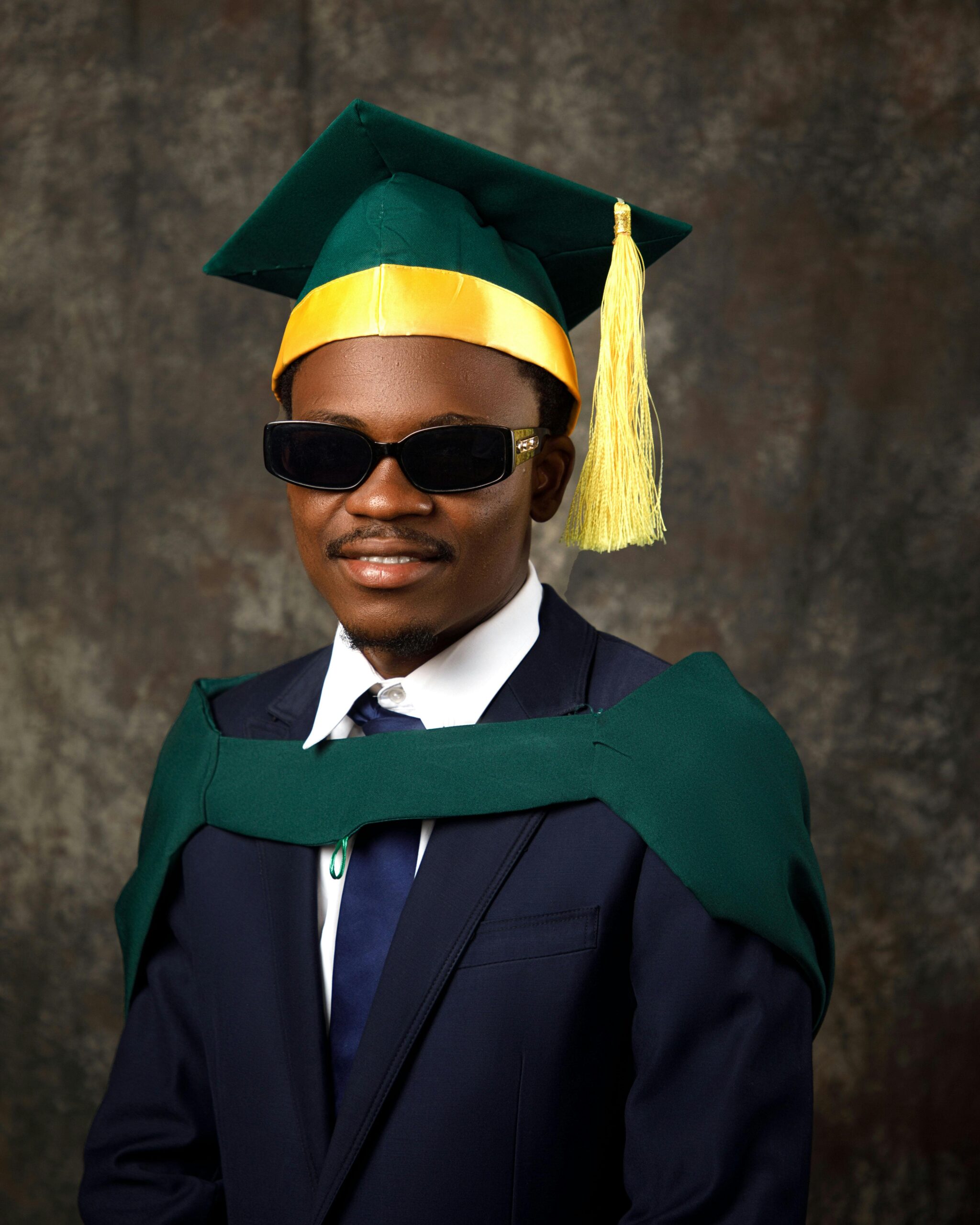Portrait of a graduate in green gown and cap, wearing sunglasses.